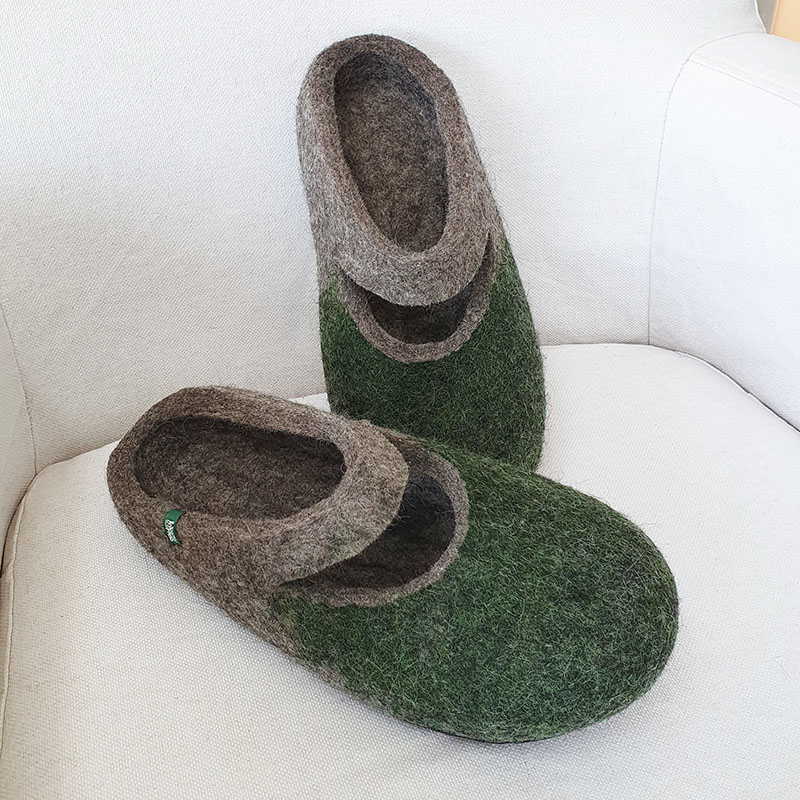 A pair of grey wool slippers with dark green on their front part. The slippers are felted without any seams. There is a cut-out piece of the slipper where the ball of the foot is, leaving an opening in the shape of a smile.