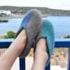 Woman's feet rested on a balcony's rail. She is wearing wool slippers in blue colors in harmony with the sea on the background. The slippers are from the SEASONS collection by Wooppers. The right slipper has turquoise on the right side of the shoe and the left has a Mediterranean blue on the left side of the shoe.