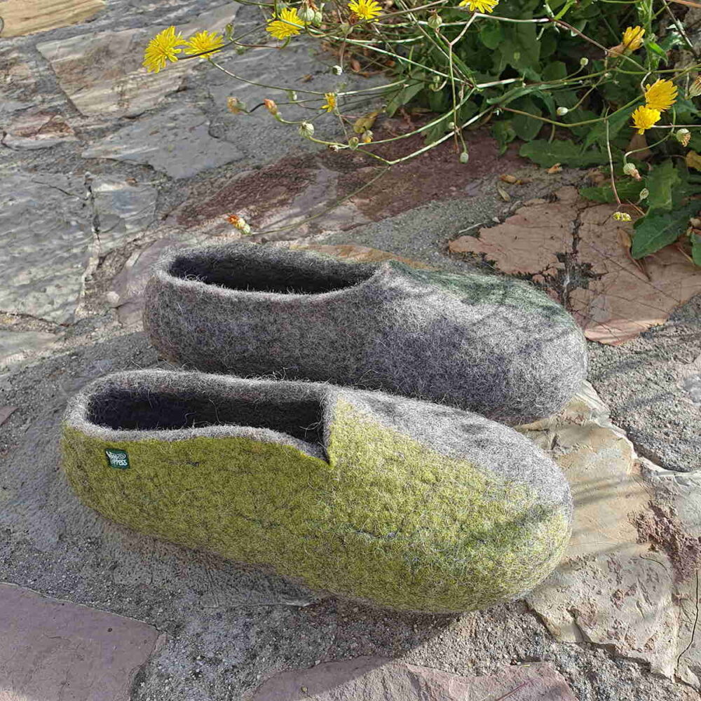 Wool slippers on a stone pathway of a garden, seen from the right side. They are slippers from the SEASONS collection by Wooppers. - grey with green colors on the sides: lime green color on the right side of the right shoe and green on the left side of the left shoe..