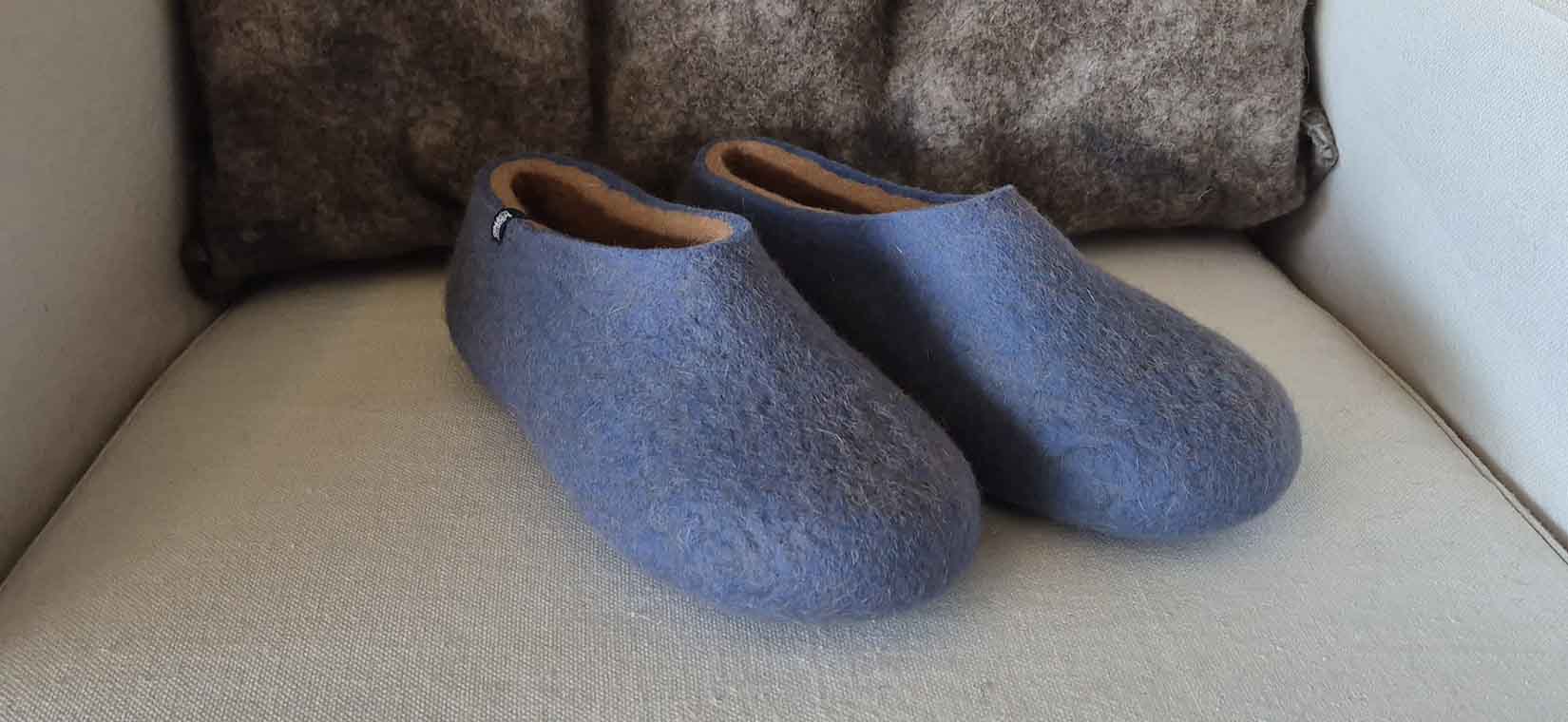 A pair of felted woolen slippers in blue and beige with a grey woolen pillow as a backdrop.